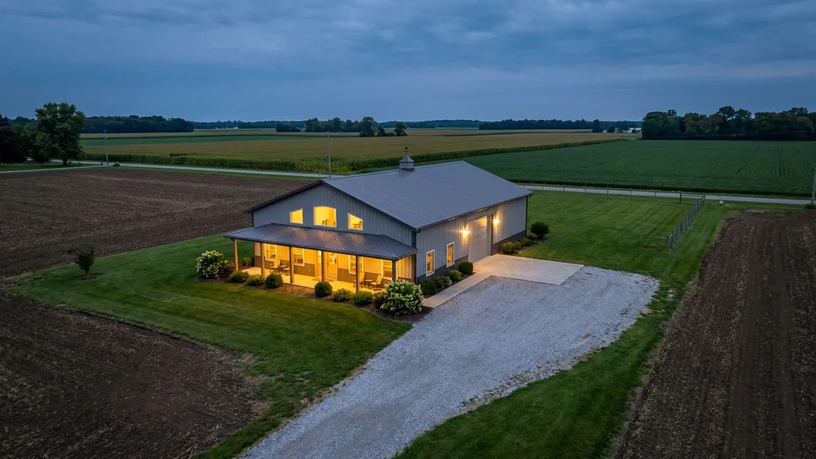 A finished barndominium at dusk with warm interior lighting glowing through its large windows.