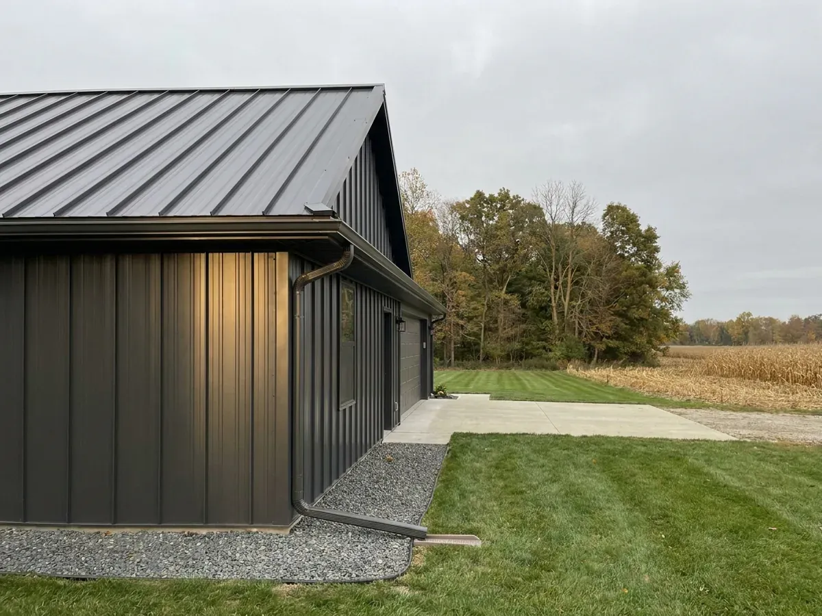 Exterior detail of a metal-clad home showing standing seam roofing, vertical wall panels, and a downspout discharging away from the wall.